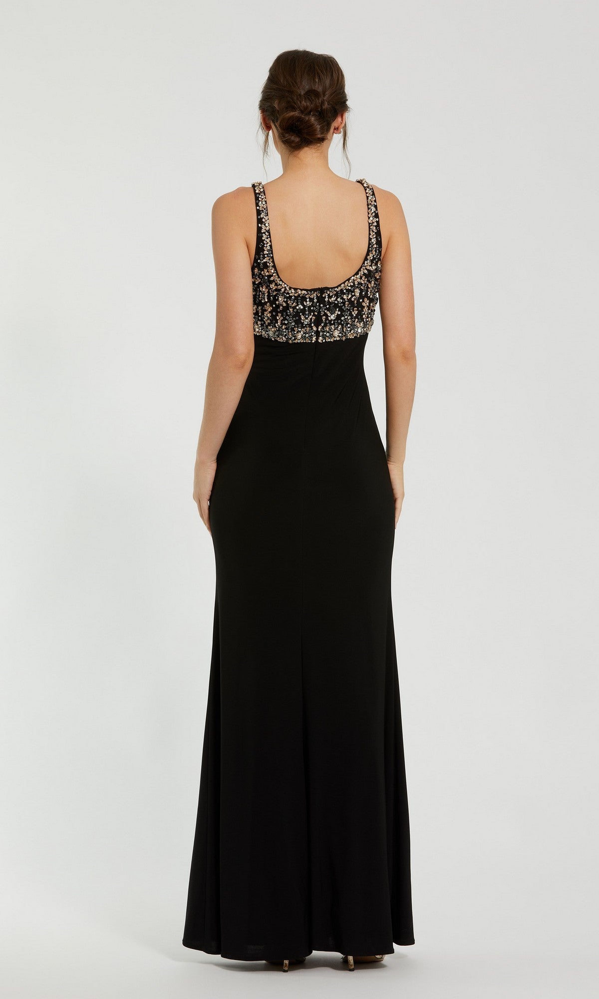 A woman stands with her back to the camera, her hair in a braided updo, wearing the Mac Duggal 2253—a long black formal dress featuring a beaded bodice and crystal embellishments—set against a plain white background; an elegant choice for evening events.